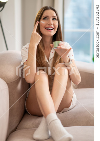 Long-haired young woman sitting on the sofa with a jar of face cream in hands Long-haired young woman sitting on the sofa with a jar of face cream in hands 137252843