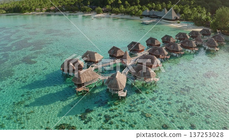 Moorea island resort featuring overwater bungalows connected by wooden walkways, sitting on a turquoise lagoon with clear shallow water, surrounded by lush green mountains Moorea island resort featuring overwater bungalows connected by wooden walkways, sitting on a turquoise lagoon with clear shallow water, surrounded by lush green mountains 137253028