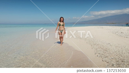 Beautiful sexy woman in green bikini walking barefoot along a pristine sandbar, surrounded by clear turquoise ocean water and distant Lombok hills under a bright blue sky, enjoying a tropical vacation 137253030