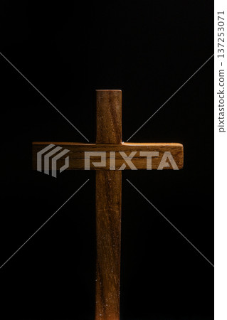 A close-up, dramatic shot of a simple wooden cross centered against a solid black background. The warm wood grain is highlighted by soft, focused lighting, symbolizing faith, hope, and spiritual 137253071