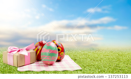 Three vibrant hand-painted Easter eggs and a gift box with a pink ribbon sit on a checkered picnic cloth in a lush green meadow under a bright blue sky. A beautiful scene of spring celebration Three vibrant hand-painted Easter eggs and a gift box with a pink ribbon sit on a checkered picnic cloth in a lush green meadow under a bright blue sky. A beautiful scene of spring celebration 137253113