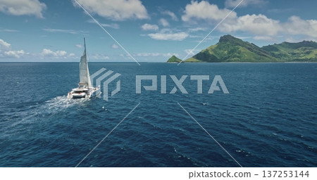 Fiji: Luxury catamaran sailing in the deep blue ocean near a tropical island with lush vegetation, aerial drone of a travel paradise. Drone flight 137253144