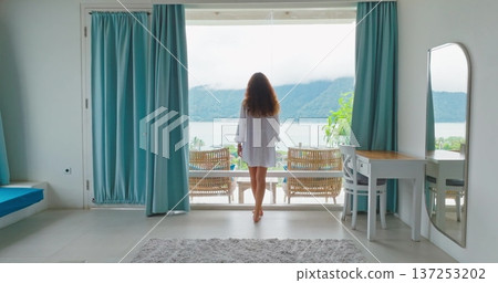 Woman in white tshirt walking hotel room balcony window, enjoy lake surrounded by lush green mountains and cloudy sky, enjoying a peaceful morning view during her tropical vacation to Bali. Back view 137253202