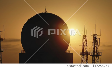 Large spherical radome and communication antenna towers silhouetted against a vibrant orange sunset sky, emphasizing the significance of telecommunications infrastructure. Aerial drone panorama 137253237