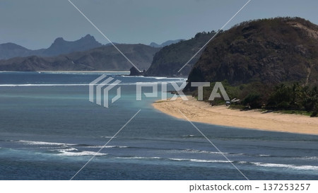 Tropical beach coastline in Lombok, Indonesia, featuring blue ocean water, sandy shore, green vegetation, and distant mountains under blue sky, an inviting travel destination. Drone aerial flight Tropical beach coastline in Lombok, Indonesia, featuring blue ocean water, sandy shore, green vegetation, and distant mountains under blue sky, an inviting travel destination. Drone aerial flight 137253257