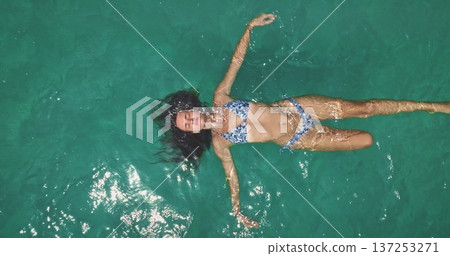 Beautiful woman in blue bikini floating on her back in crystal clear turquoise ocean water, enjoying a relaxing travel vacation and summer holiday recreation. Top down aerial view zoom out Beautiful woman in blue bikini floating on her back in crystal clear turquoise ocean water, enjoying a relaxing travel vacation and summer holiday recreation. Top down aerial view zoom out 137253271