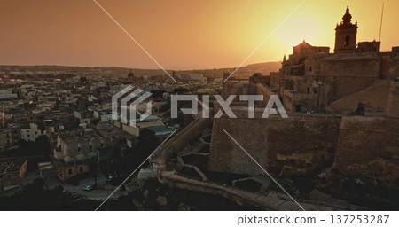 Malta, Comino island: Warm sunset light illuminating the Cittadella bastions in Gozo, Malta, blending history with a modern urban landscape. Drone flight over evening city 137253287
