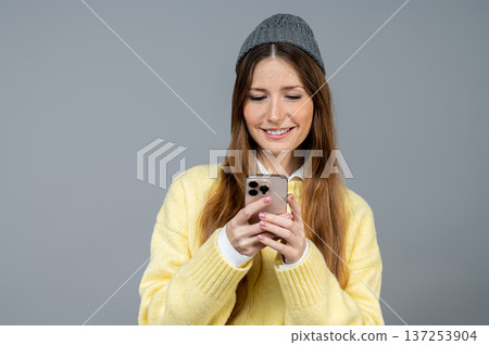 Smiling and contented young woman in grey hat texting on her phone and feeling good Smiling and contented young woman in grey hat texting on her phone and feeling good 137253904