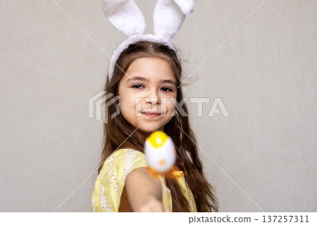 Girl wearing bunny ears offers a decorated Easter egg with a playful smile during spring celebration 137257311