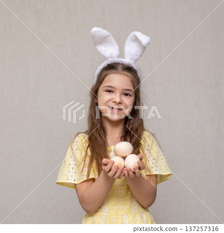 Smiling girl in bunny ears holding wooden eggs in yellow dress for Easter celebration Smiling girl in bunny ears holding wooden eggs in yellow dress for Easter celebration 137257316