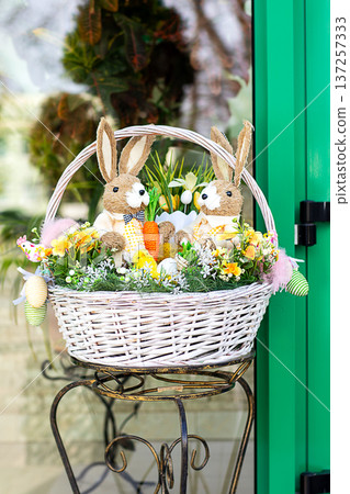 Easter basket with two plush bunny toys, flowers and decorative eggs on a metal stand Easter basket with two plush bunny toys, flowers and decorative eggs on a metal stand 137257333