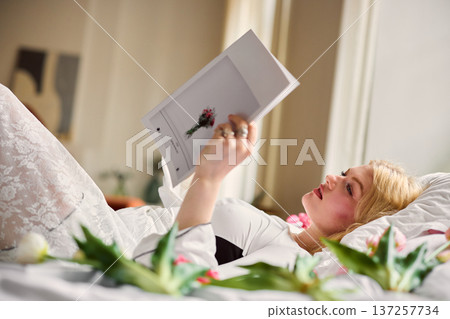 Casually reclined woman on white fabric observes vibrant floral editorial content with focused interest Casually reclined woman on white fabric observes vibrant floral editorial content with focused interest 137257734
