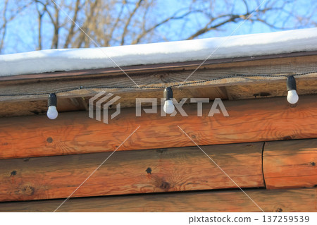 Rustic log cabin wall with a snow-covered roof and a hanging garland of light bulbs in winter. Rustic log cabin wall with a snow-covered roof and a hanging garland of light bulbs in winter. 137259539