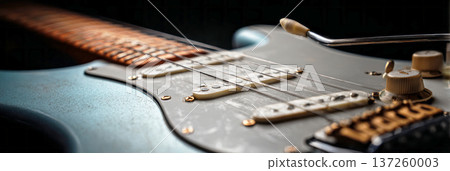 A guitar rests on a wooden table with soft light highlighting its body and strings. Knobs and switches catch the light. The room has a warm atmosphere as music fills the air, banner 137260003