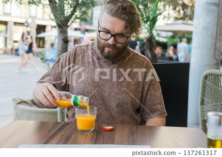 Man pouring orange juice into glass while sitting at outdoor cafe table. Urban lifestyle, casual refreshment ritual, simple pleasure, and everyday beverage moment during relaxed city leisure 137261317