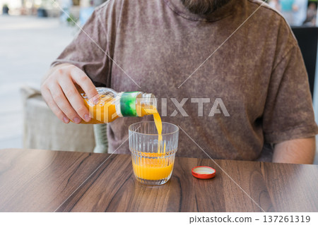 Fresh orange juice pouring from bottle into glass at cafe terrace table. Beverage preparation, summer refreshment, thirst relief, and relaxed urban lifestyle moment in outdoor dining setting Fresh orange juice pouring from bottle into glass at cafe terrace table. Beverage preparation, summer refreshment, thirst relief, and relaxed urban lifestyle moment in outdoor dining setting 137261319