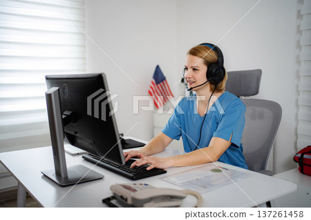 Healthcare worker wearing scrubs and headset smiling while typing on computer 137261458