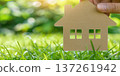 Hand holding a house cutout over lush green grass in a bright, sunlit garden setting 137261942