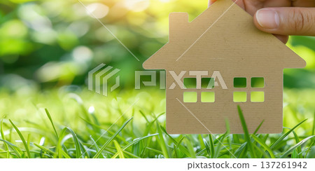 Hand holding a house cutout over lush green grass in a bright, sunlit garden setting 137261942