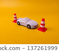 Toy sports car positioned next to three miniature red and white traffic cones on a yellow surface. 137261999