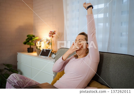 Tired woman yawning and stretching while sitting on sofa with laptop 137262011