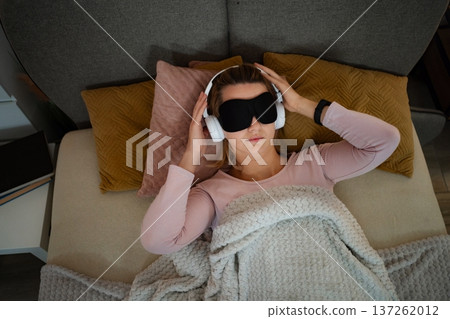 Woman relaxing in bed listening to music wearing sleep mask 137262012