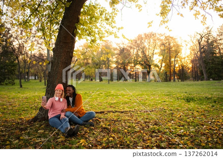 Diverse friends capturing selfie sitting under autumn tree in park Diverse friends capturing selfie sitting under autumn tree in park 137262014