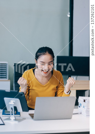 Asian woman works on laptop at office desk. She smiles happily while typing on keyboard near window with blinds. Professional woman works in business environment. Asian woman works on laptop at office desk. She smiles happily while typing on keyboard near window with blinds. Professional woman works in business environment. 137262101