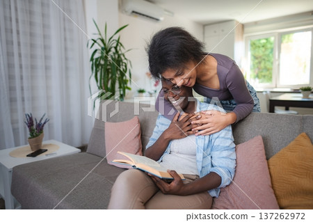 Young Woman Embracing Her Mother Reading a Book on the Sofa at Home Young Woman Embracing Her Mother Reading a Book on the Sofa at Home 137262972