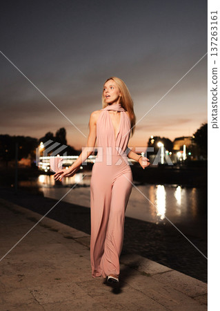 Elegant woman strolls by the river at sunset in soft pink attire 137263161