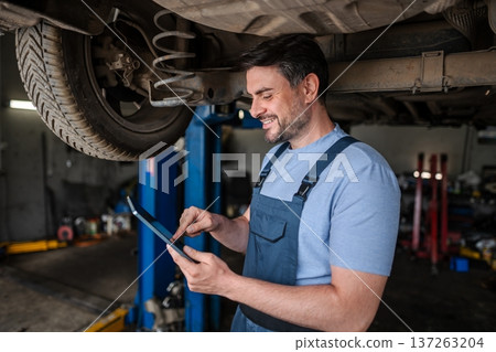 Car mechanic using a digital tablet while working under a lifted 137263204