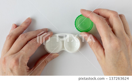 Hands holding contact lenses case white background, closeup. Woman hands unscrew the green lid of a white contact lens container Hands holding contact lenses case white background, closeup. Woman hands unscrew the green lid of a white contact lens container 137263624
