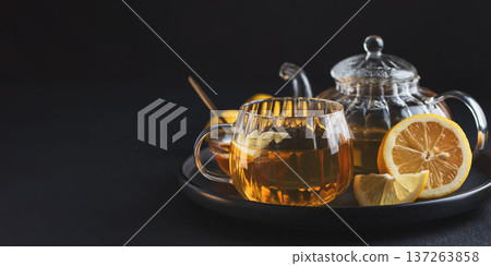 Glass teapot with amber tea, teacup with lemon slice, and bowl of honey with golden spoon on dark concrete background 137263858