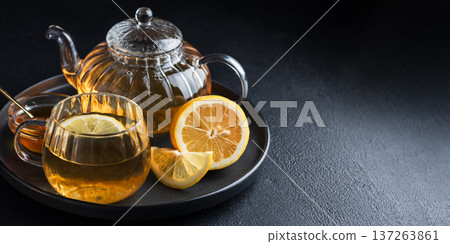 Glass teapot with amber tea, teacup with lemon slice, and bowl of honey with golden spoon on dark concrete background 137263861