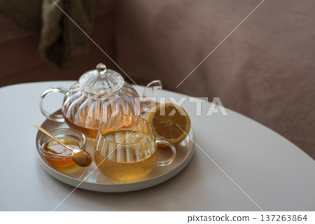 Glass teapot with amber tea, teacup with lemon slice, and bowl of honey with golden spoon on white table in cozy living room 137263864