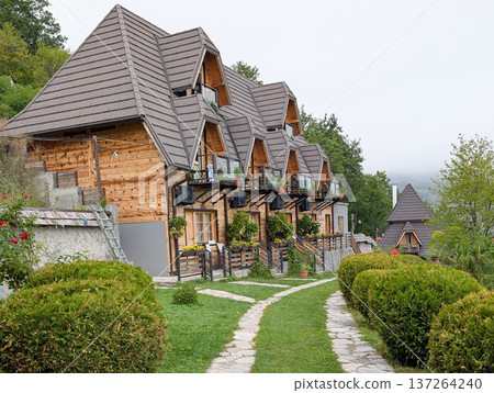 Row of small wooden cottages with pointed roofs, surrounded by trimmed bushes and stone path. Tourism and mountain accommodation in Serbia. Row of small wooden cottages with pointed roofs, surrounded by trimmed bushes and stone path. Tourism and mountain accommodation in Serbia. 137264240