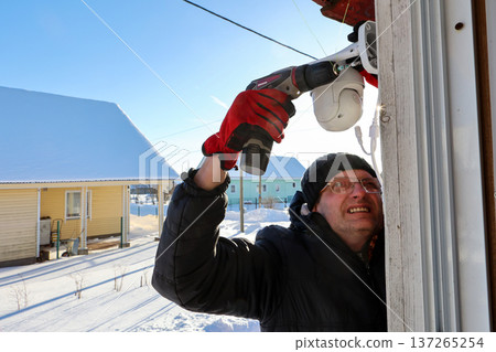 Outdoor dome surveillance camera mounting on facade of rural house, farmer tightens fastener using screwdriver. 137265254