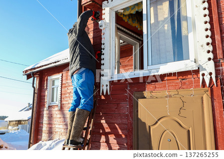 Farmer mounts dome surveillance camera on window casing of country house, standing on stepladder. 137265255