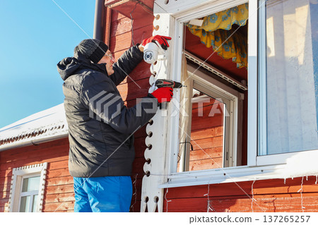 Outdoor surveillance camera mounting on facade of farm house in winter, farmer stands on stepladder holding screwdriver in hands. Outdoor surveillance camera mounting on facade of farm house in winter, farmer stands on stepladder holding screwdriver in hands. 137265257
