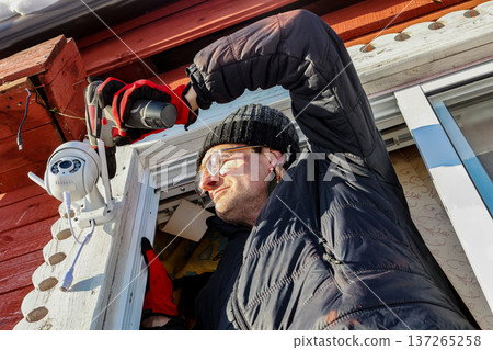 Installer tightens mounting screws of outdoor surveillance camera using screwdriver. 137265258