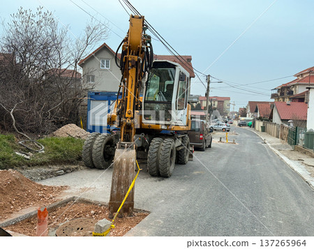 Excavator working on road repair in a suburban residential street with houses and utility lines. Urban infrastructure maintenance, municipal construction activity, civil engineering and public service 137265964