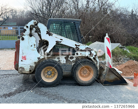 Compact skid steer loader parked on asphalt near roadwork barriers and construction materials. Urban infrastructure maintenance, heavy equipment operation and municipal engineering activity. 137265966