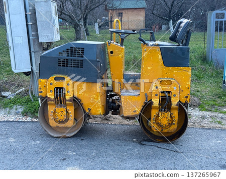 Yellow road roller parked near freshly paved asphalt in residential street during infrastructure maintenance. Transportation modernization, civil engineering process and municipal road improvement Yellow road roller parked near freshly paved asphalt in residential street during infrastructure maintenance. Transportation modernization, civil engineering process and municipal road improvement 137265967