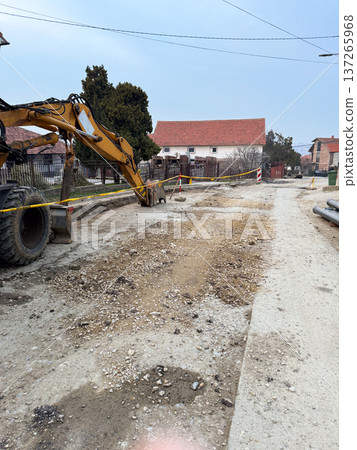 Excavator digging trench during road reconstruction in suburban neighborhood with safety tape and construction barriers. Urban infrastructure repair, civil engineering, transportation maintenance and Excavator digging trench during road reconstruction in suburban neighborhood with safety tape and construction barriers. Urban infrastructure repair, civil engineering, transportation maintenance and 137265968