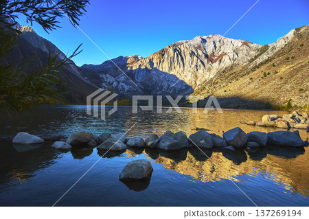 Mountain Lake Reflections at Sunrise with Rocky Shoreline and Clear Blue Sky 137269194
