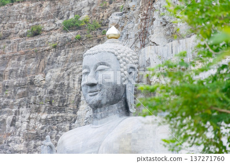 Large stone Buddha statue carved against rocky cliff face with serene expression and golden ushnisha spiritual landmark surrounded by natural landscape Large stone Buddha statue carved against rocky cliff face with serene expression and golden ushnisha spiritual landmark surrounded by natural landscape 137271760