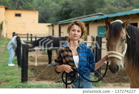 Happy woman farmer in plaid shirt and gloves preparing horse for riding and walking in pasture on farm 137274227