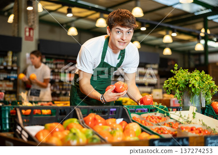 Cheerful guy seller arranging ripe tomatoes on counter in grocery store 137274253