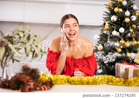 Emotional young female in red dress talking on mobile phone close to packed gift boxes and decorated Christmas tree at apartment Emotional young female in red dress talking on mobile phone close to packed gift boxes and decorated Christmas tree at apartment 137274295