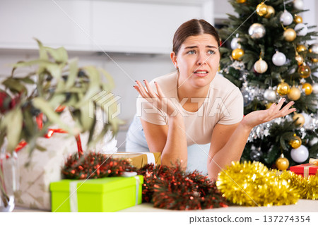 Emotional sad young female next to packed gift box with bow near and decorated Christmas tree at home 137274354